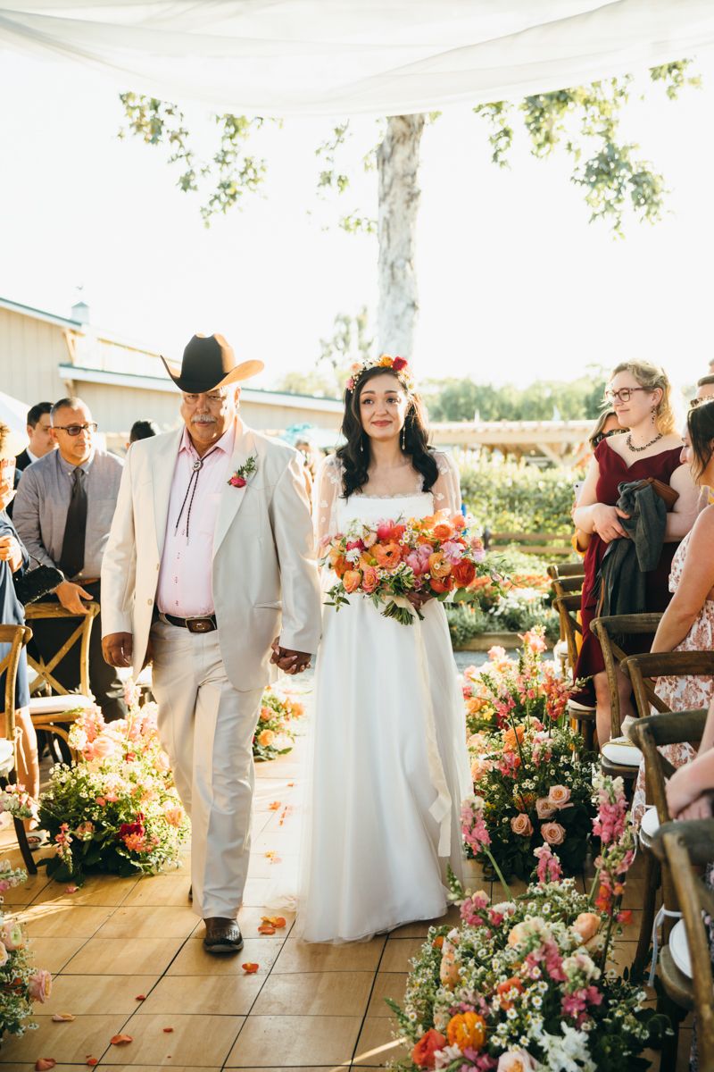 Father walks bride down floral-lined aisle at outdoor wedding ceremony.