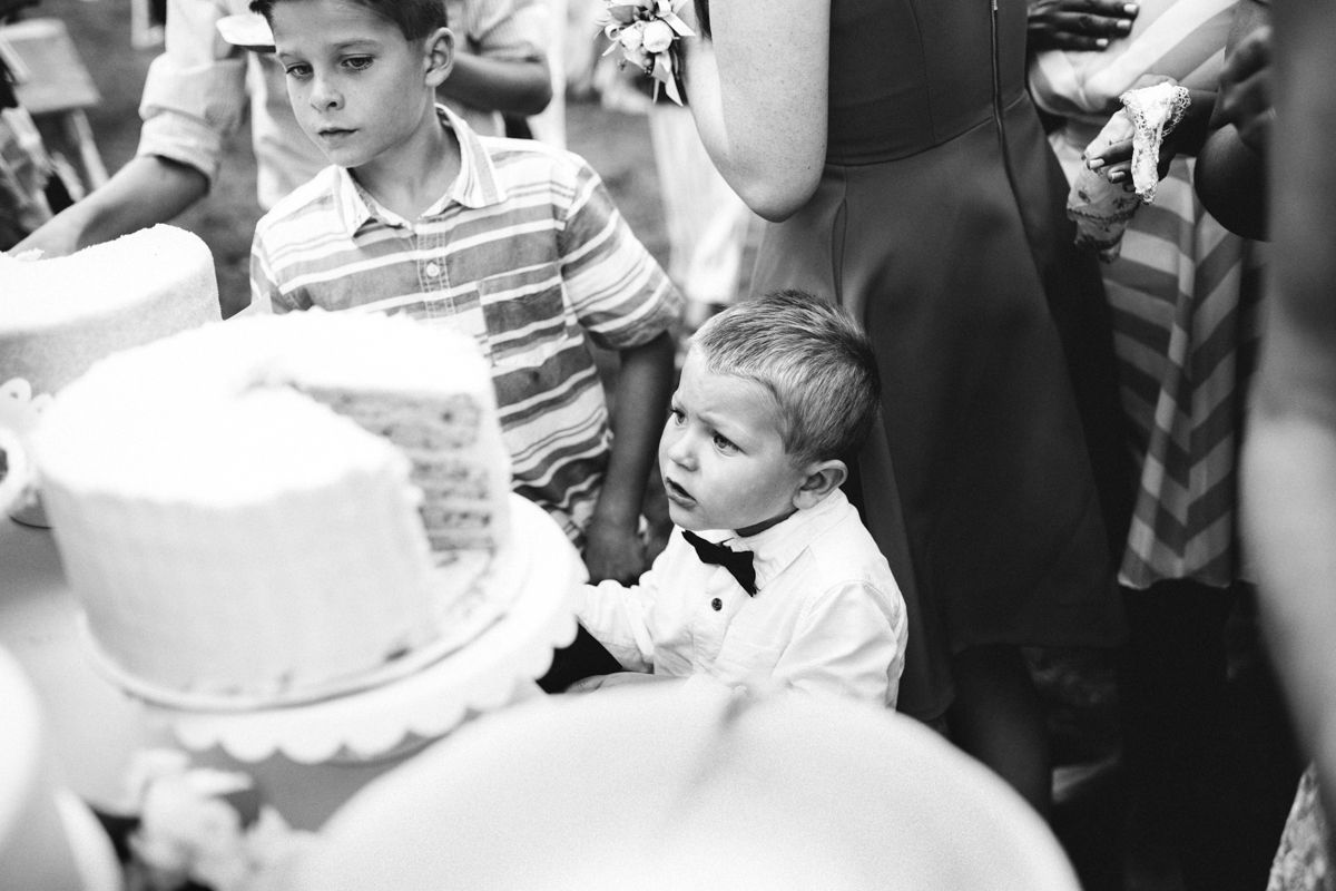 A young boy in a bow tie gazes at a white wedding cake in a black and white photo.