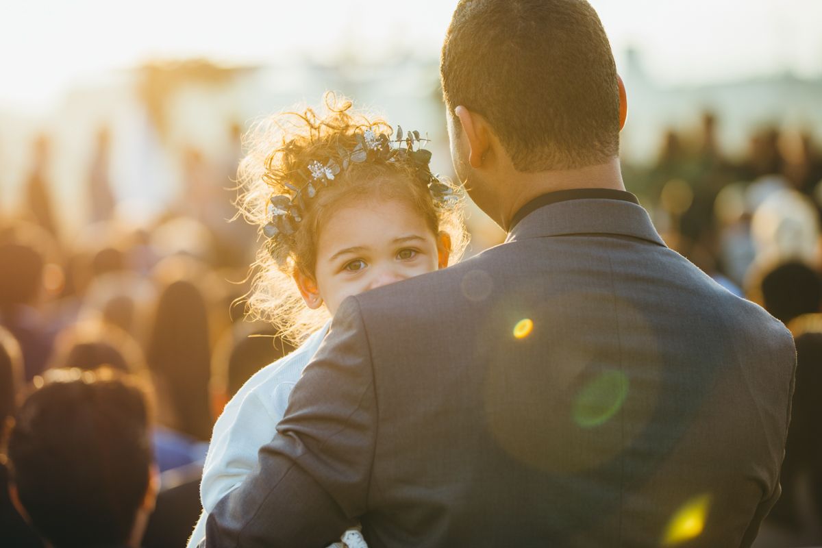 A man in a suit holds a curly-haired little girl against a golden sunset at a wedding.