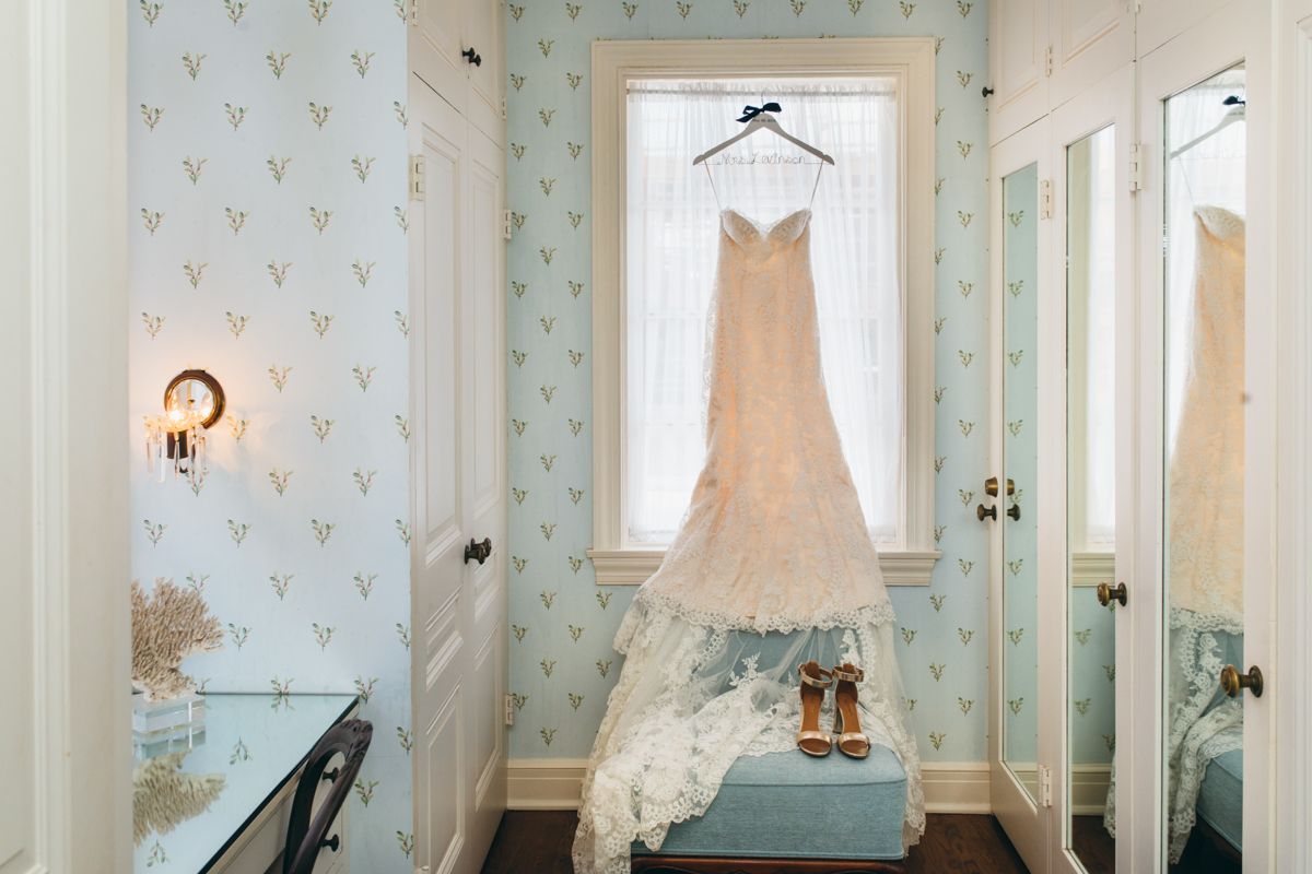 Wedding dress hanging in window of elegant bridal suite with blue patterned wallpaper and vintage decor.