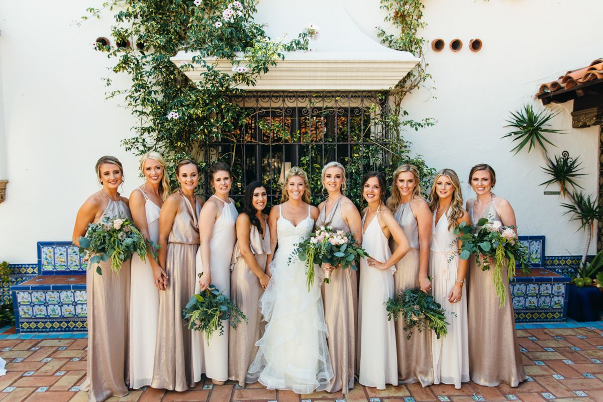 Bride surrounded by bridesmaids in champagne gowns holding lush greenery bouquets outdoors.