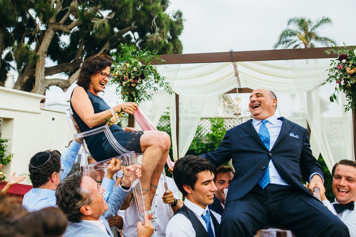 Jewish wedding hora dance with bride and groom lifted in chairs by joyful guests outdoors.
