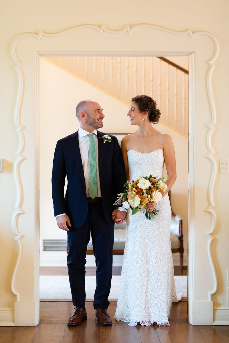 Bride and groom share a romantic moment standing in an ornate white doorway, bride holding autumn bouquet.