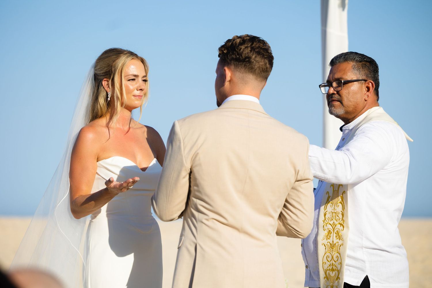 Bride and groom exchange vows at a beach wedding ceremony with an officiant under a clear blue sky.
