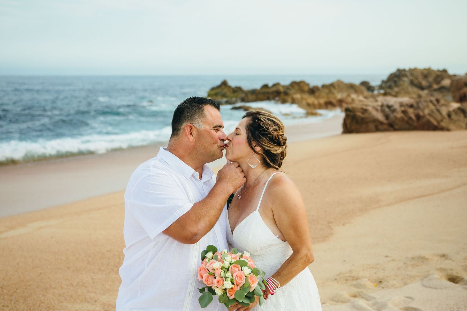 Couple sharing a romantic kiss on a sandy beach in cabo, bride holding a peach bouquet with ocean waves and rocks in the background.