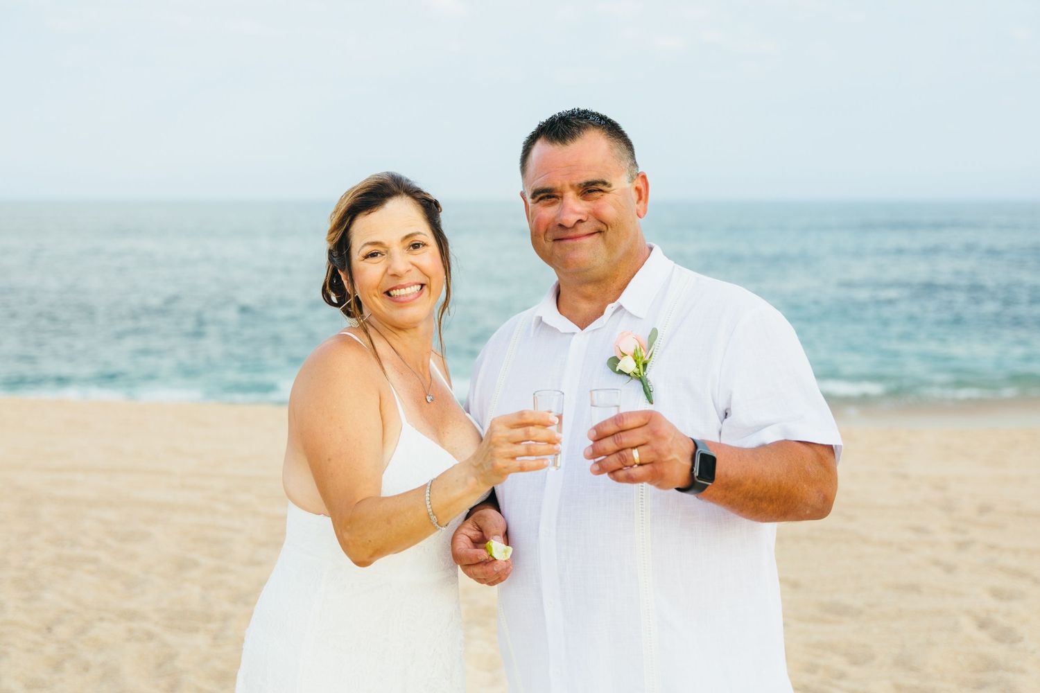 Couple in white attire celebrating on a sandy beach, holding champagne glasses with ocean waves in the background.