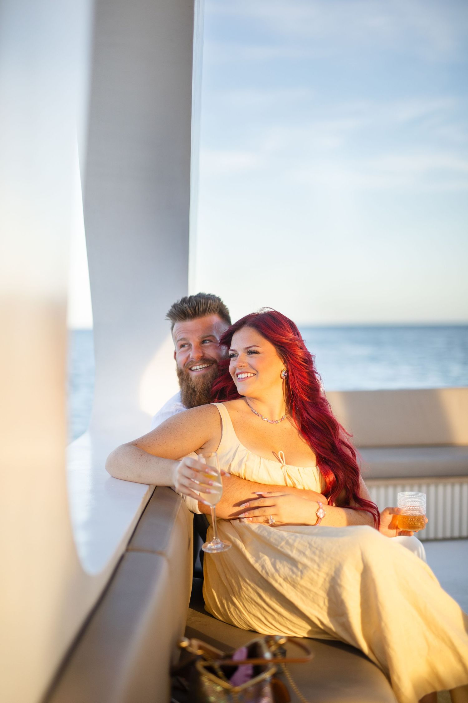 Joyful couple embracing on a luxury yacht at sunset, woman with red hair holding a glass of wine.