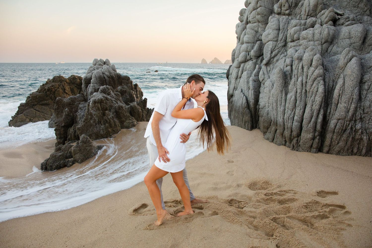 Couple sharing a romantic kiss on a rocky beach at sunset in Cabo San Lucas.