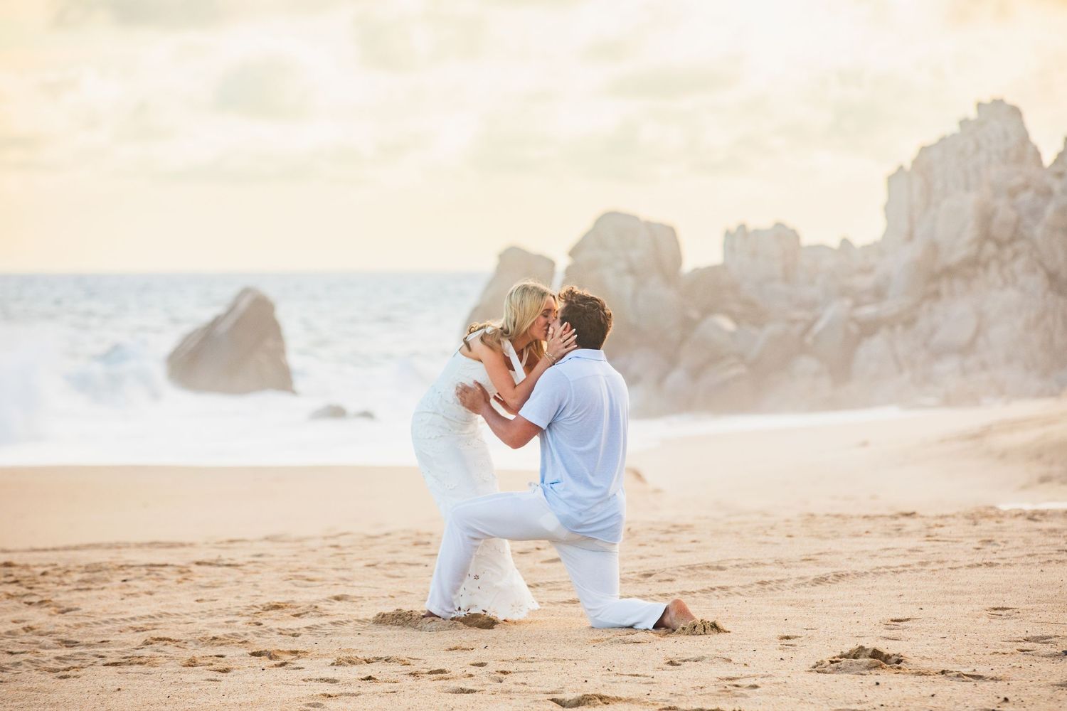 Couple sharing a kiss on the beach after proposal, man still on one knee in romantic moment.
