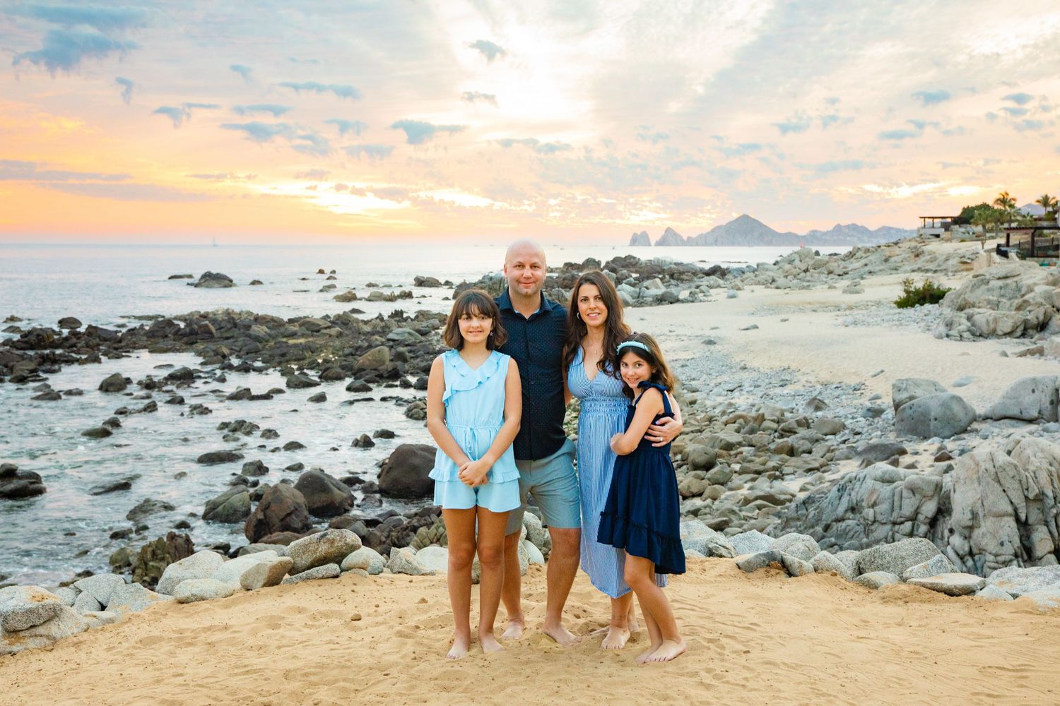 Family of four posing on a rocky beach at sunset in Cabo San Lucas, wearing coordinating blue outfits.