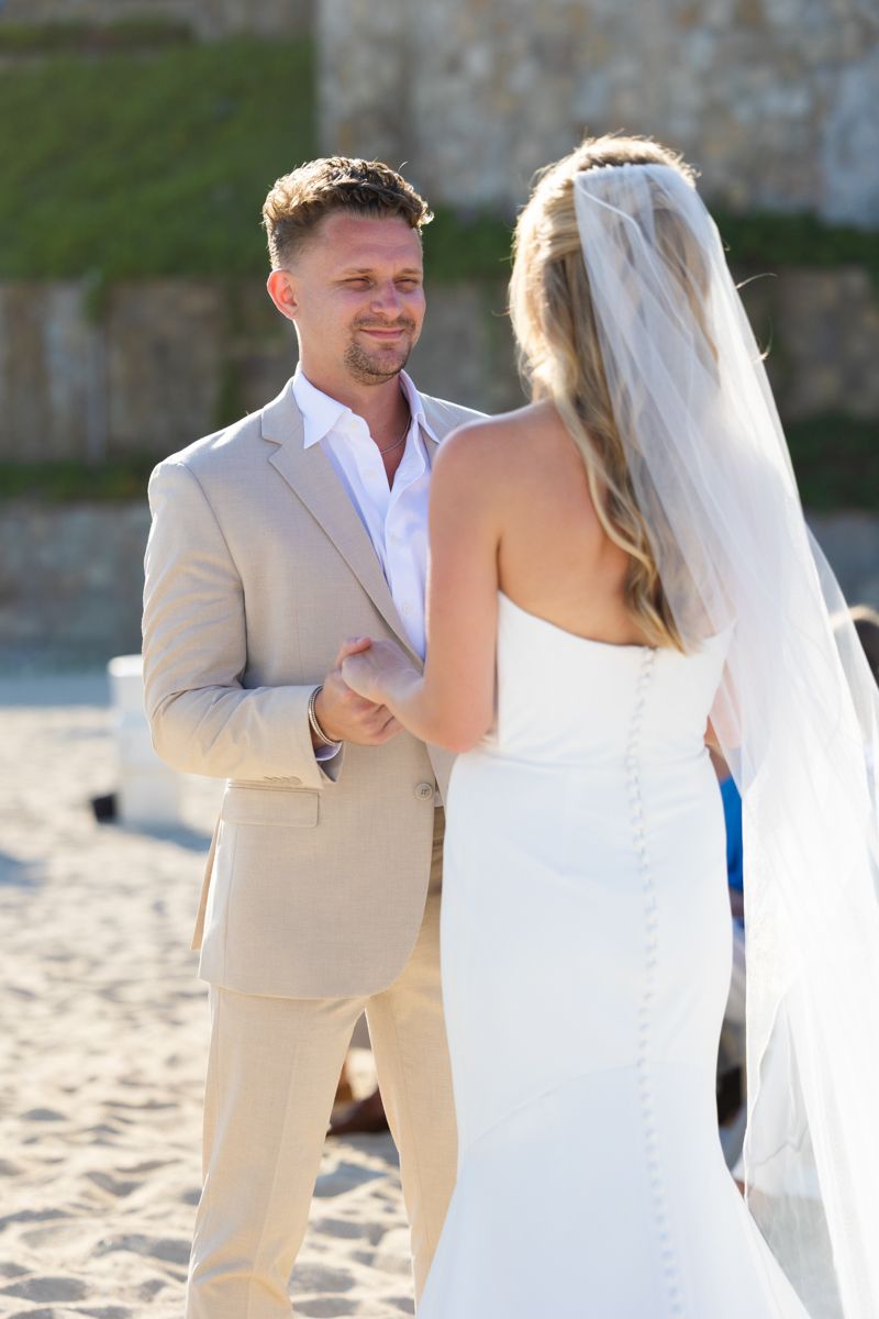 Groom smiles tenderly at bride during outdoor beach wedding ceremony vow exchange.