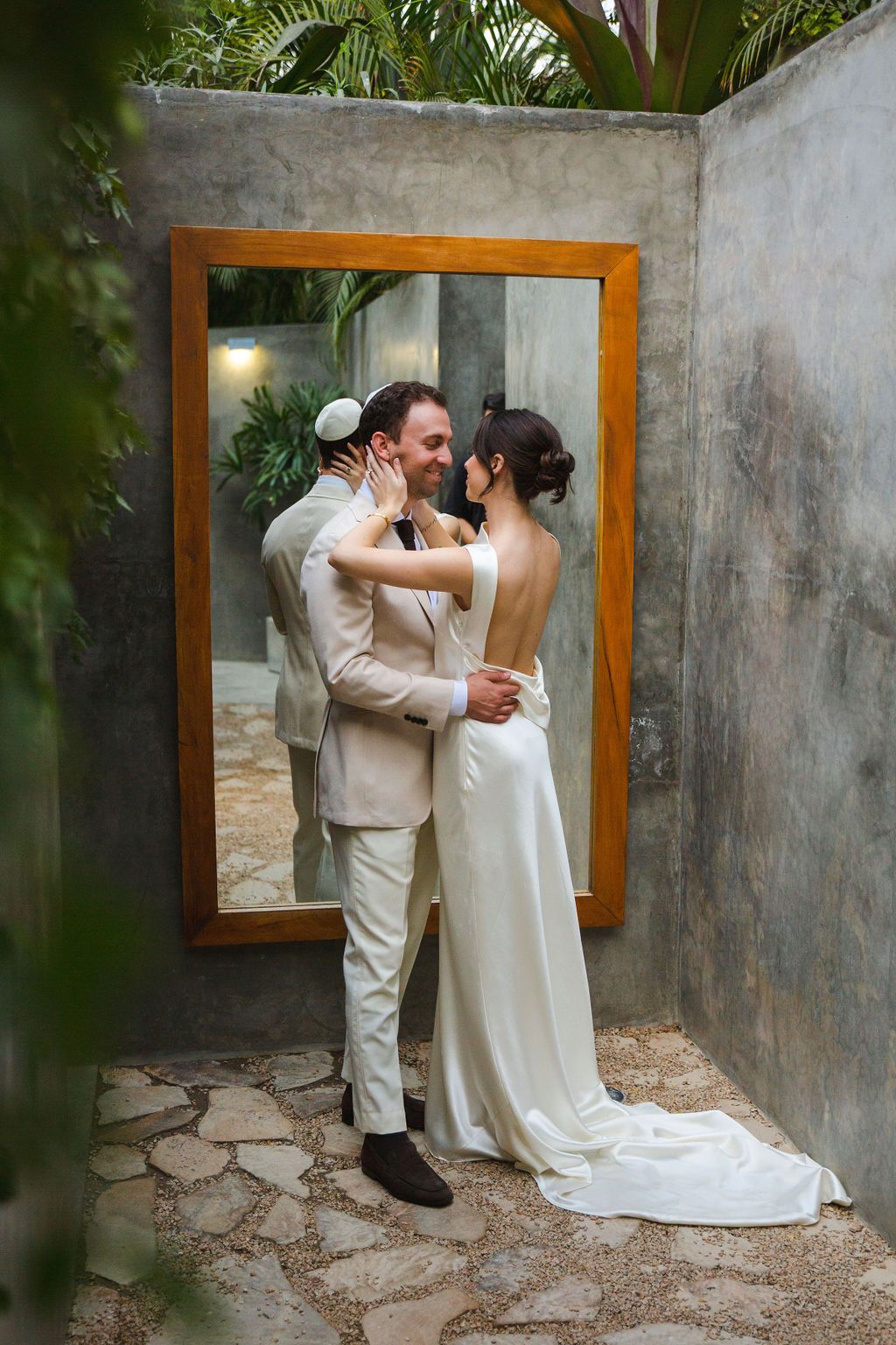Bride and groom sharing a romantic embrace in front of a wooden mirror in a tropical garden setting.