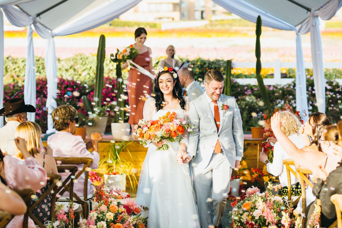 Bride and groom walk down flower-lined aisle at colorful outdoor wedding ceremony.