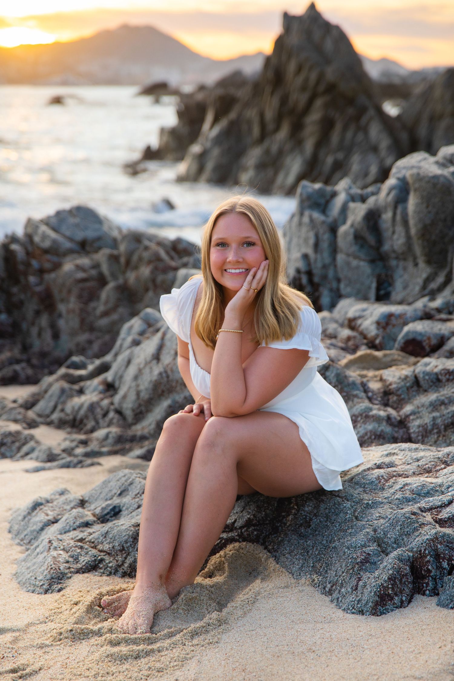 White sundress on coastal rocks during golden sunset with waves and rugged shoreline in background.