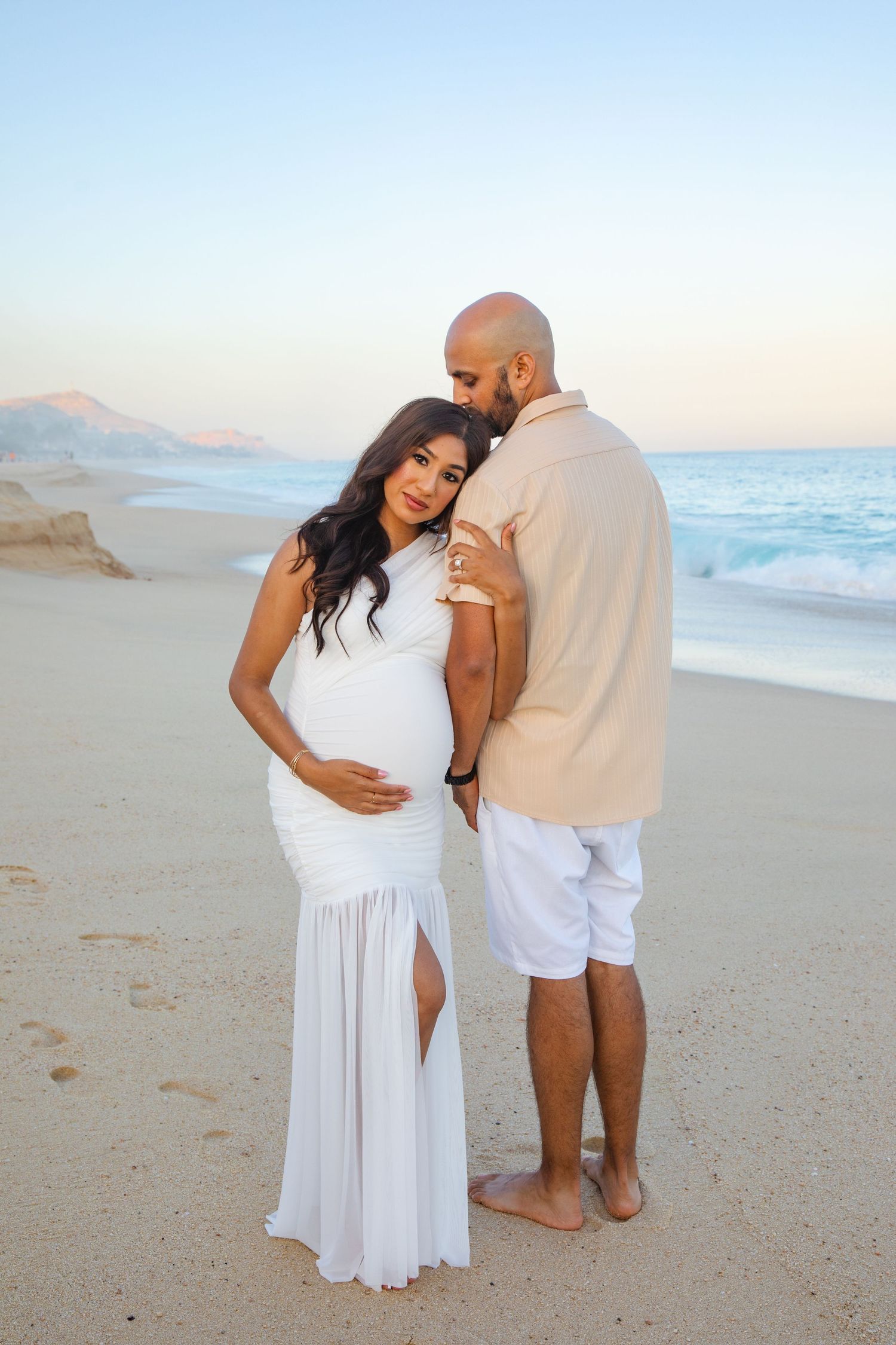 Pregnant couple embracing on a beach in Cabo San Lucas at sunset, woman in white maternity gown, man in beige shirt and white shorts.