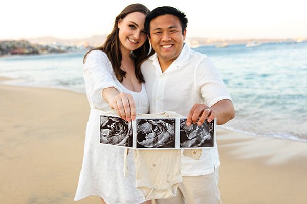 Happy couple on beach holding ultrasound photos and baby onesie announcing pregnancy.