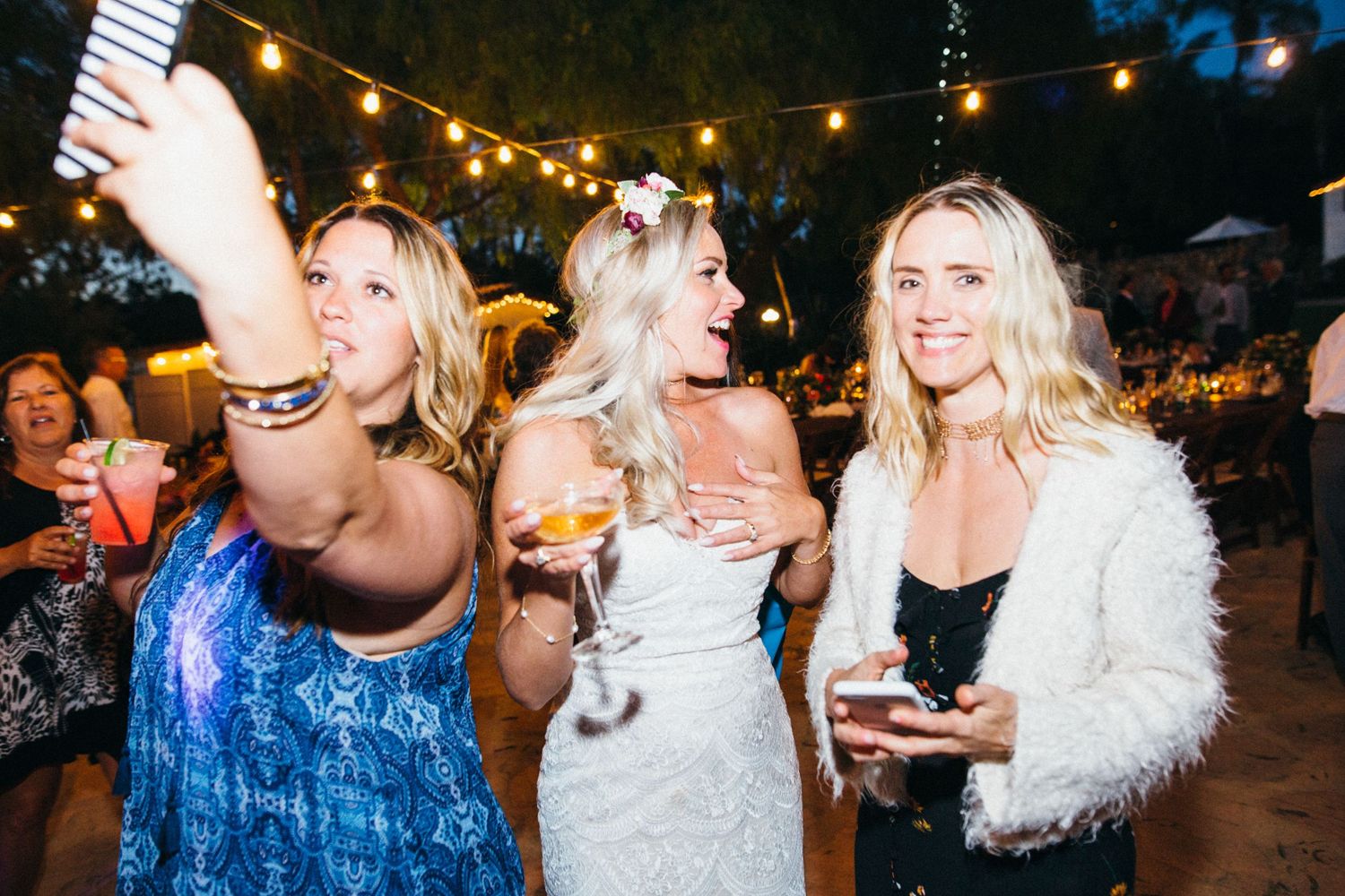 Three women smiling and taking a selfie at an outdoor evening party with string lights.
