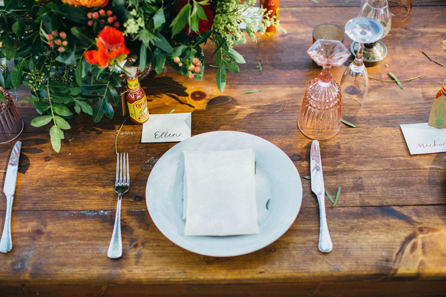 Rustic wooden table setting with white plate, folded napkin, silverware, pink glassware, and vibrant orange floral centerpiece.