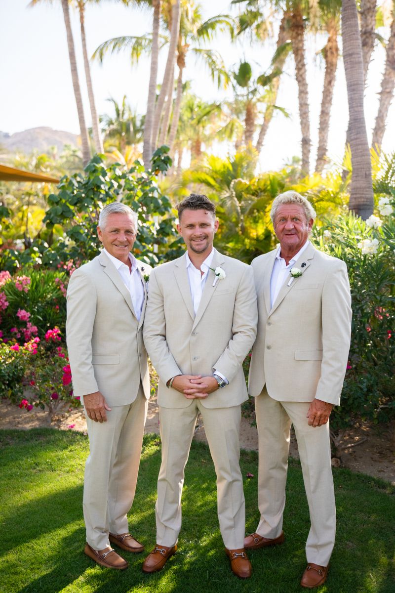 Groom poses with two groomsmen in matching tan suits among tropical palm tree setting.