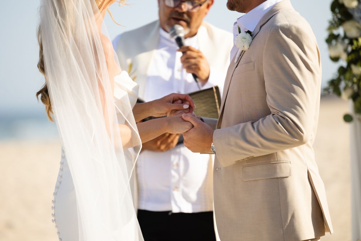 Groom placing wedding ring on bride's finger during beach ceremony with officiant in background.