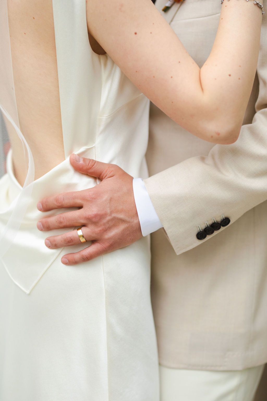 Close-up of groom in beige suit with gold wedding band embracing bride in white satin wedding gown.