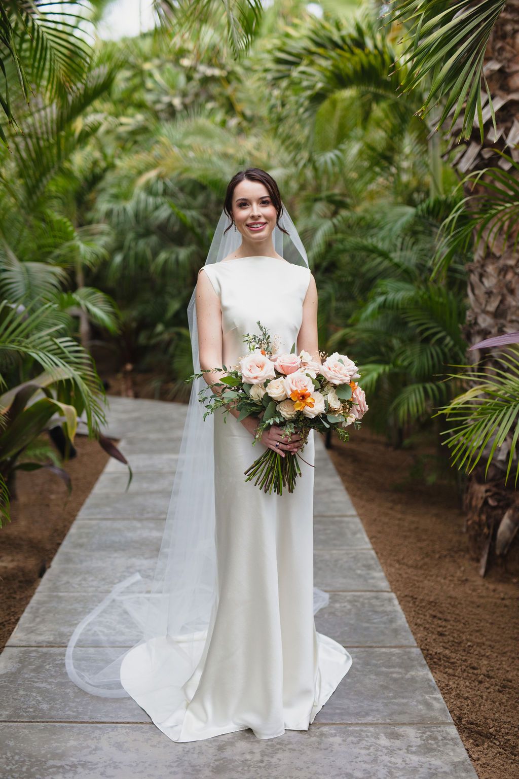 Smiling bride in sleek white gown and veil holds peach and white bouquet in tropical garden setting.