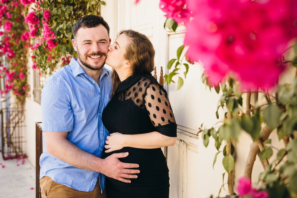 Happy couple embracing near pink bougainvillea, woman kissing mans cheek outdoors.