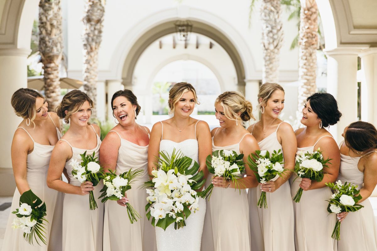 A bride and seven bridesmaids in champagne gowns hold white floral bouquets under elegant arches.