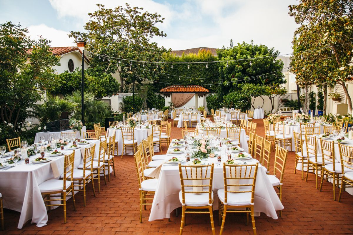 Outdoor wedding reception setup with white tablecloths, gold chiavari chairs, and floral centerpieces on a brick patio.