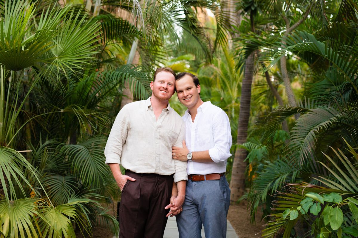 Two men embracing and smiling together surrounded by lush tropical palm trees and greenery.