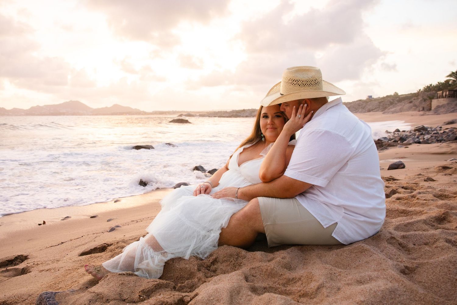 Couple sits romantically on a sandy beach at sunset, woman wearing a straw hat.