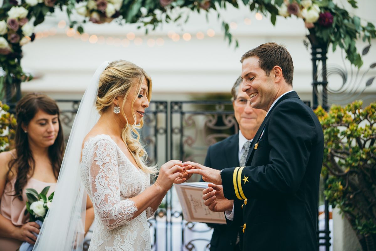 Bride places wedding ring on groom's finger during outdoor ceremony, groom smiling in navy uniform.