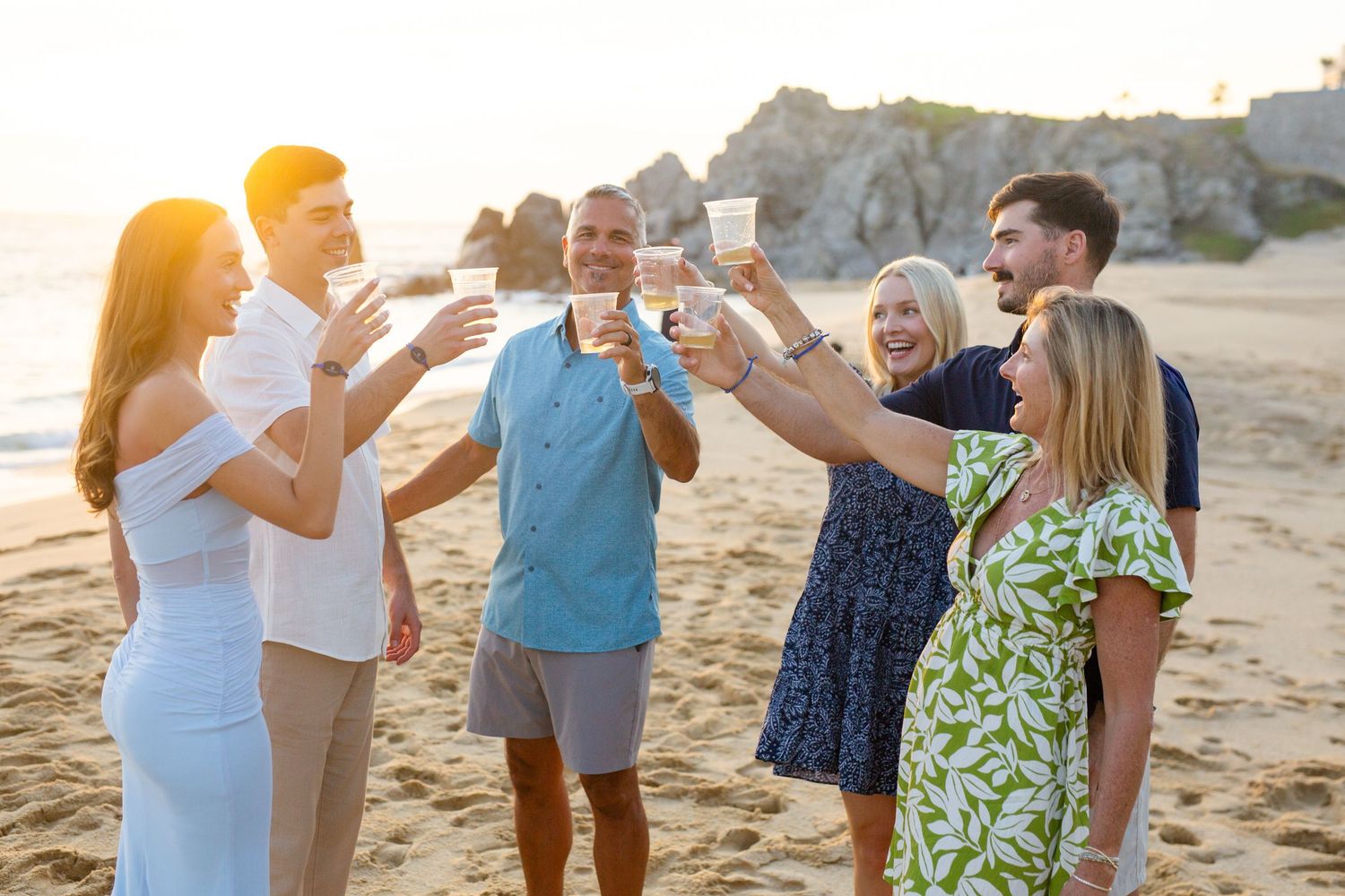 Group of friends toasting drinks on a sunny beach at sunset, celebrating together with smiles and raised glasses in cabo