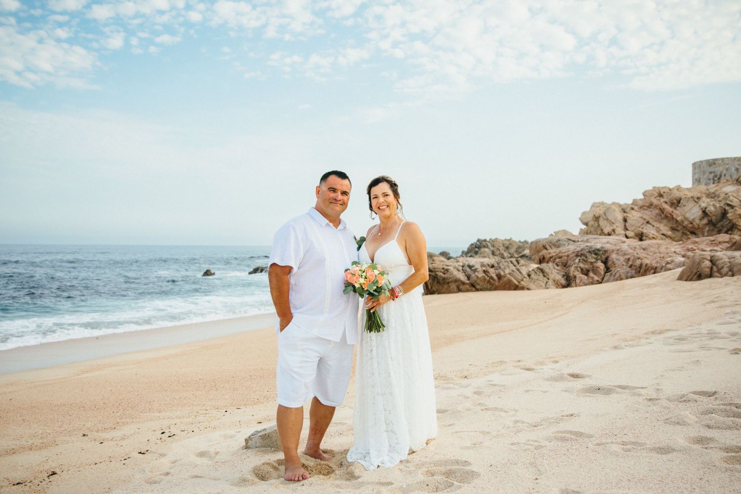 Couple in white attire poses on sandy beach in cabo with ocean waves and rocky cliffs during outdoor wedding ceremony.