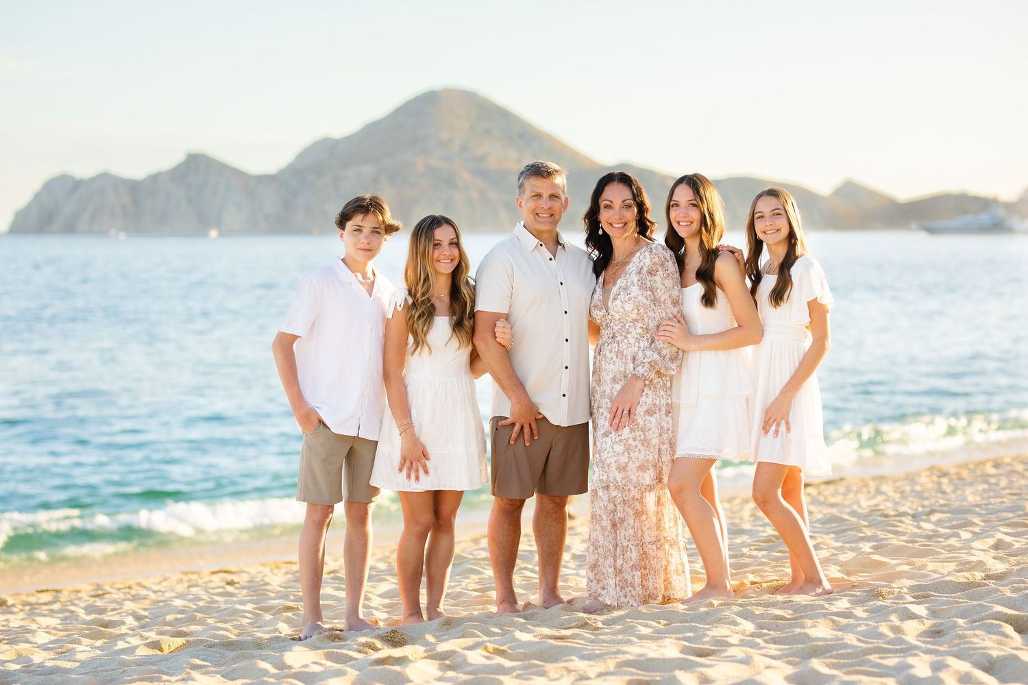 Happy family of six posing together on a sandy beach with a mountain and ocean backdrop.