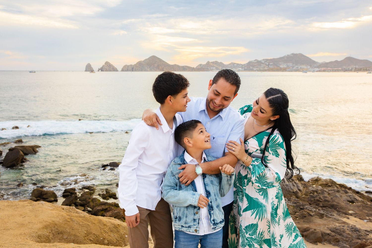 Joyful family of four laughing together on rocky coastal overlook at sunset.