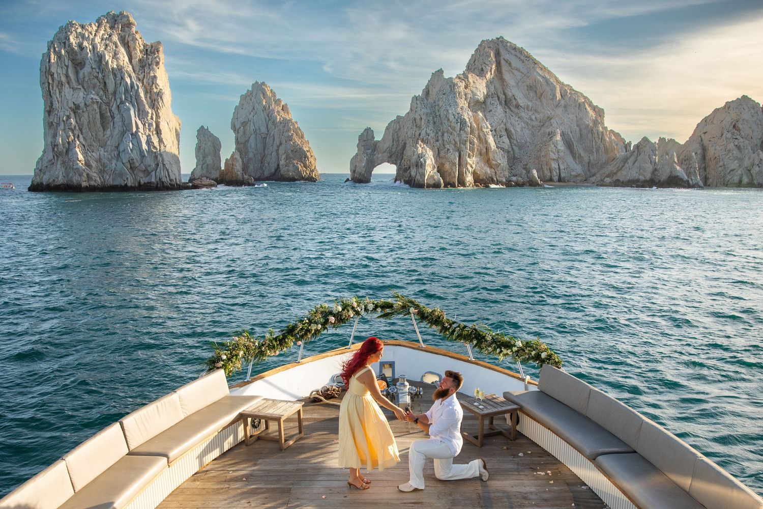 Man proposes to woman on luxury yacht bow near iconic Arch of Cabo San Lucas rock formation in Mexico.