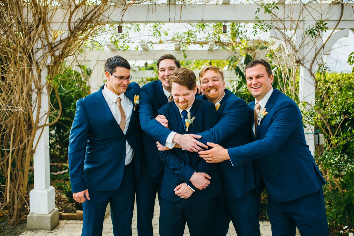 Groom and groomsmen in navy suits laugh together outdoors near white pergola.