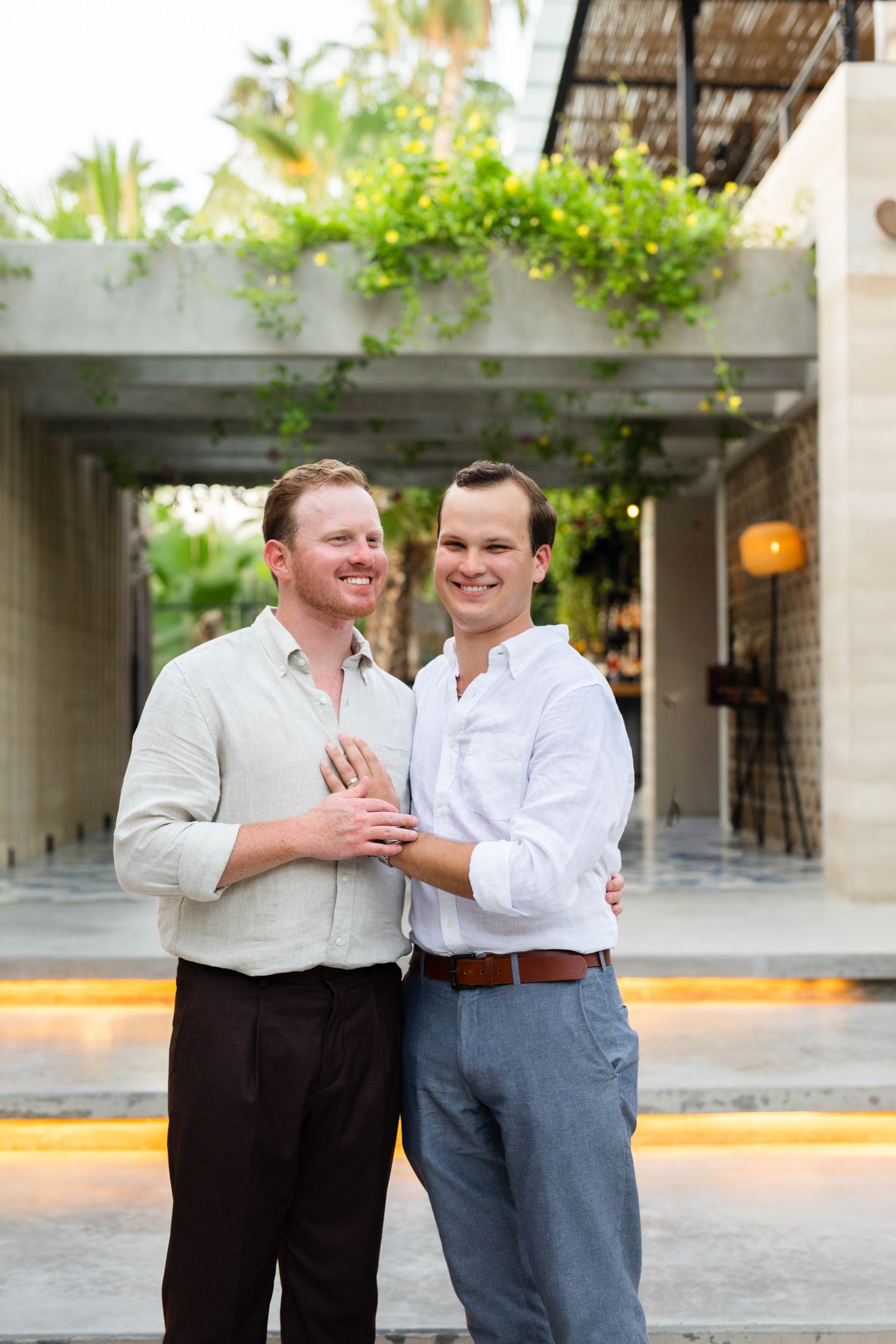 Two smiling men embrace outdoors at a modern venue with lush greenery, dressed in casual button-up shirts.