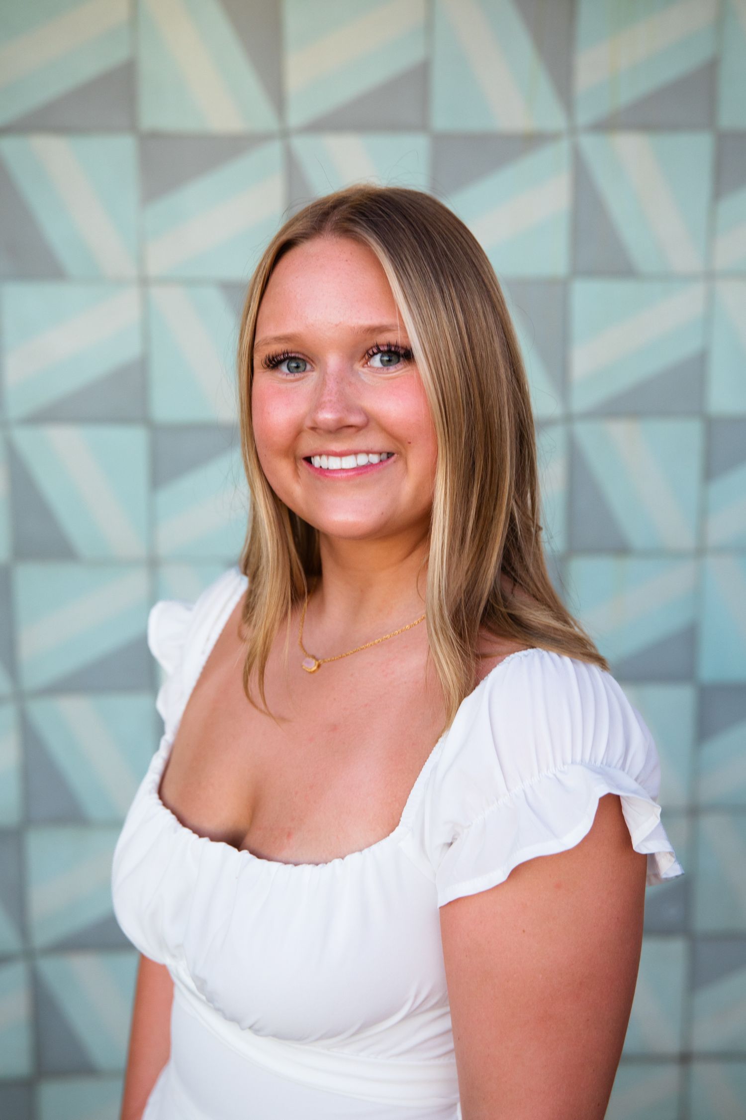 Smiling blonde woman in white ruffled top posing in front of a geometric teal patterned wall.