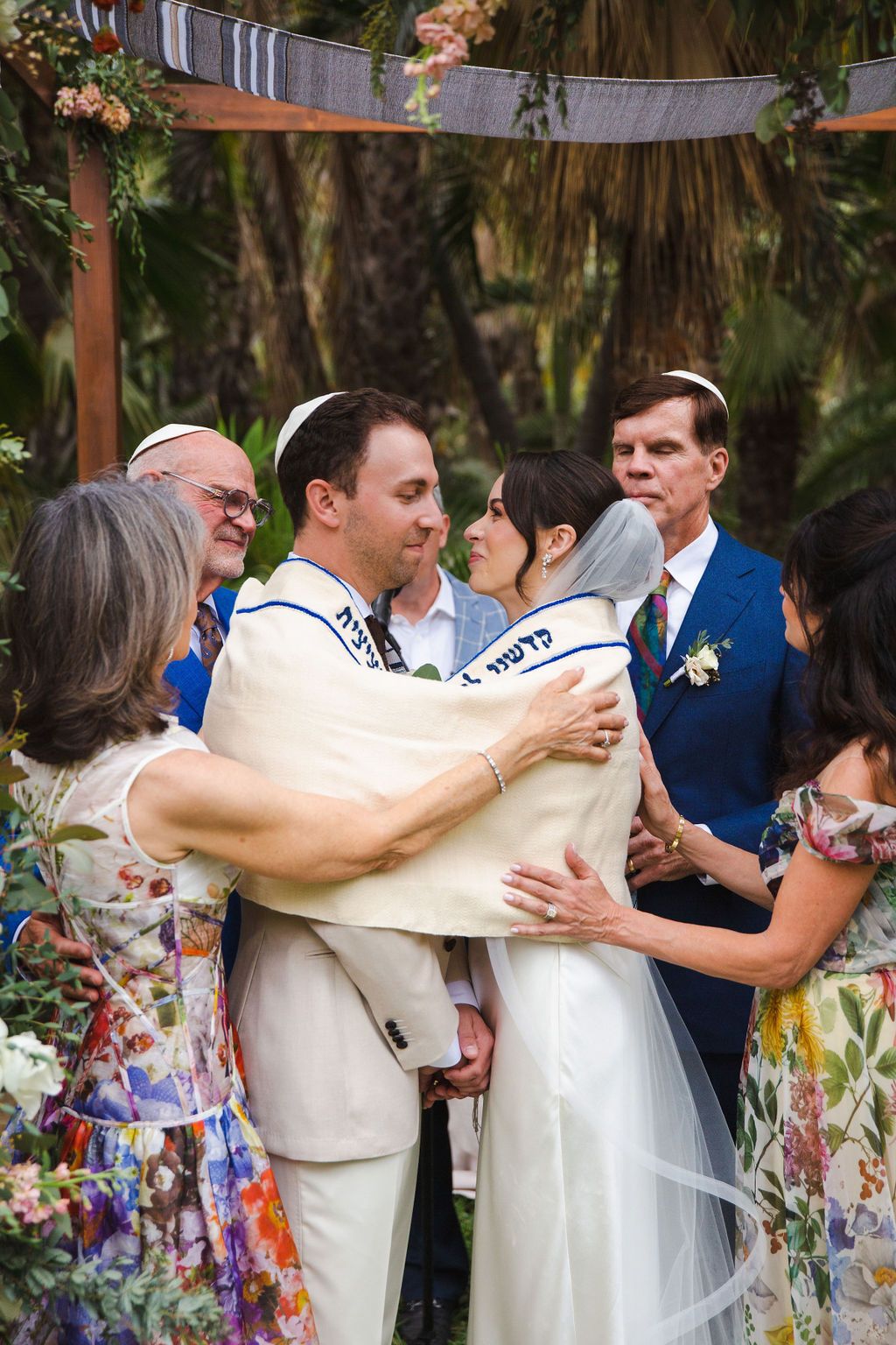 Bride and groom sharing a warm embrace with family during a Jewish wedding ceremony under a floral chuppah.