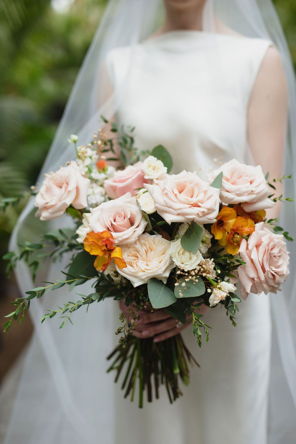 Bride holding lush wedding bouquet of blush pink roses and orange flowers with greenery.