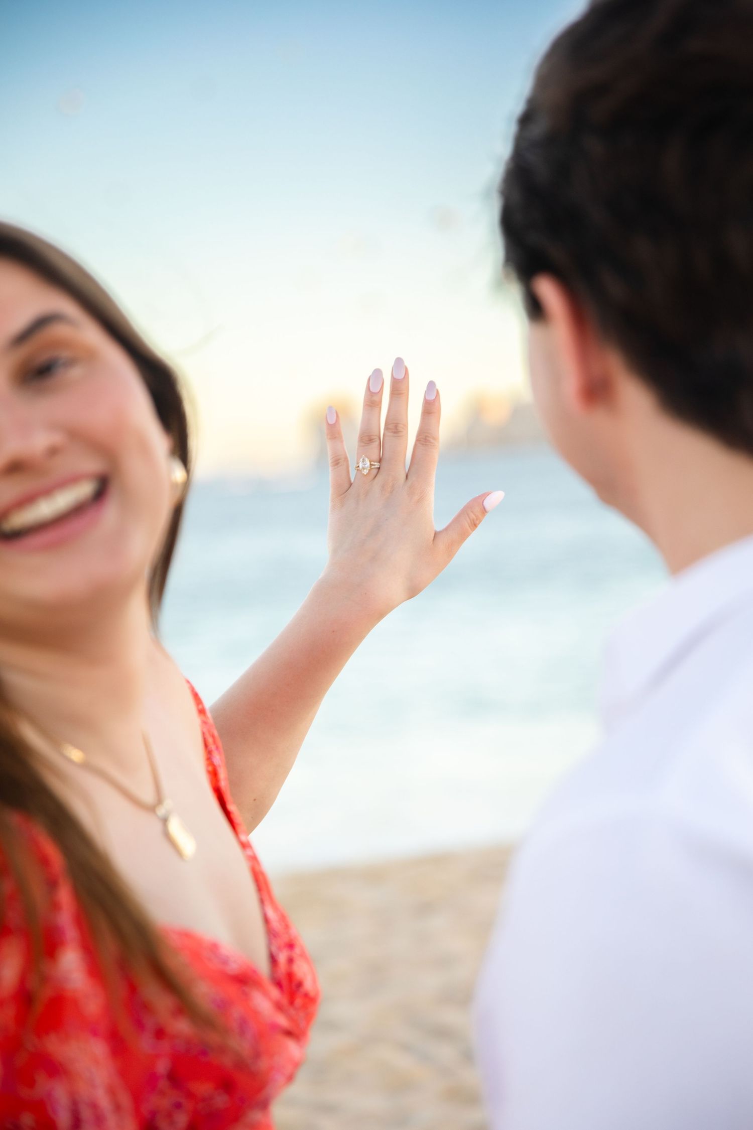 Woman in red dress smiling on beach showing off engagement ring to partner at sunset.