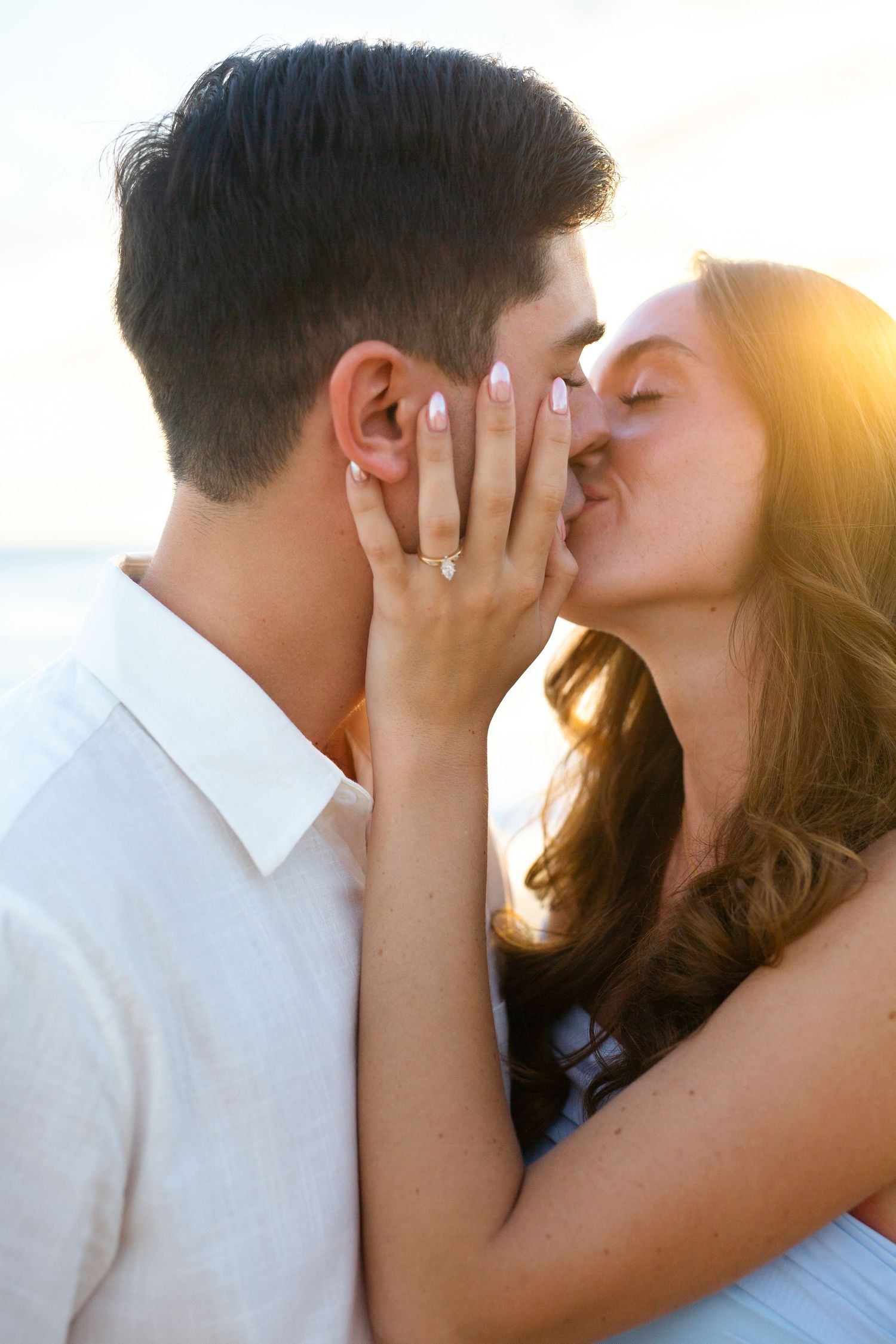 Couple sharing a romantic kiss at sunset on the beach, woman displaying engagement ring on mans cheek.
