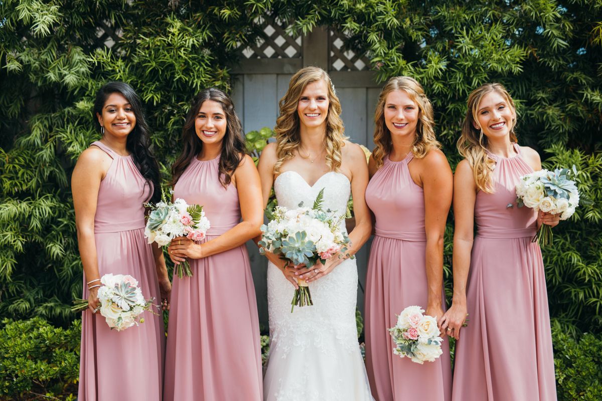Bride in lace gown poses with four bridesmaids in dusty rose dresses holding white bouquets.