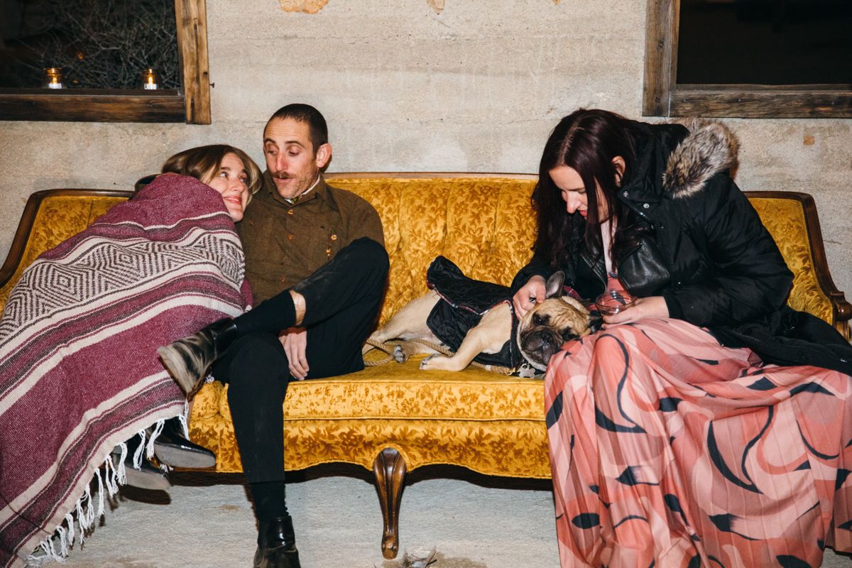 Three tired wedding guests relax on a golden velvet sofa with a small dog at the end of the night.