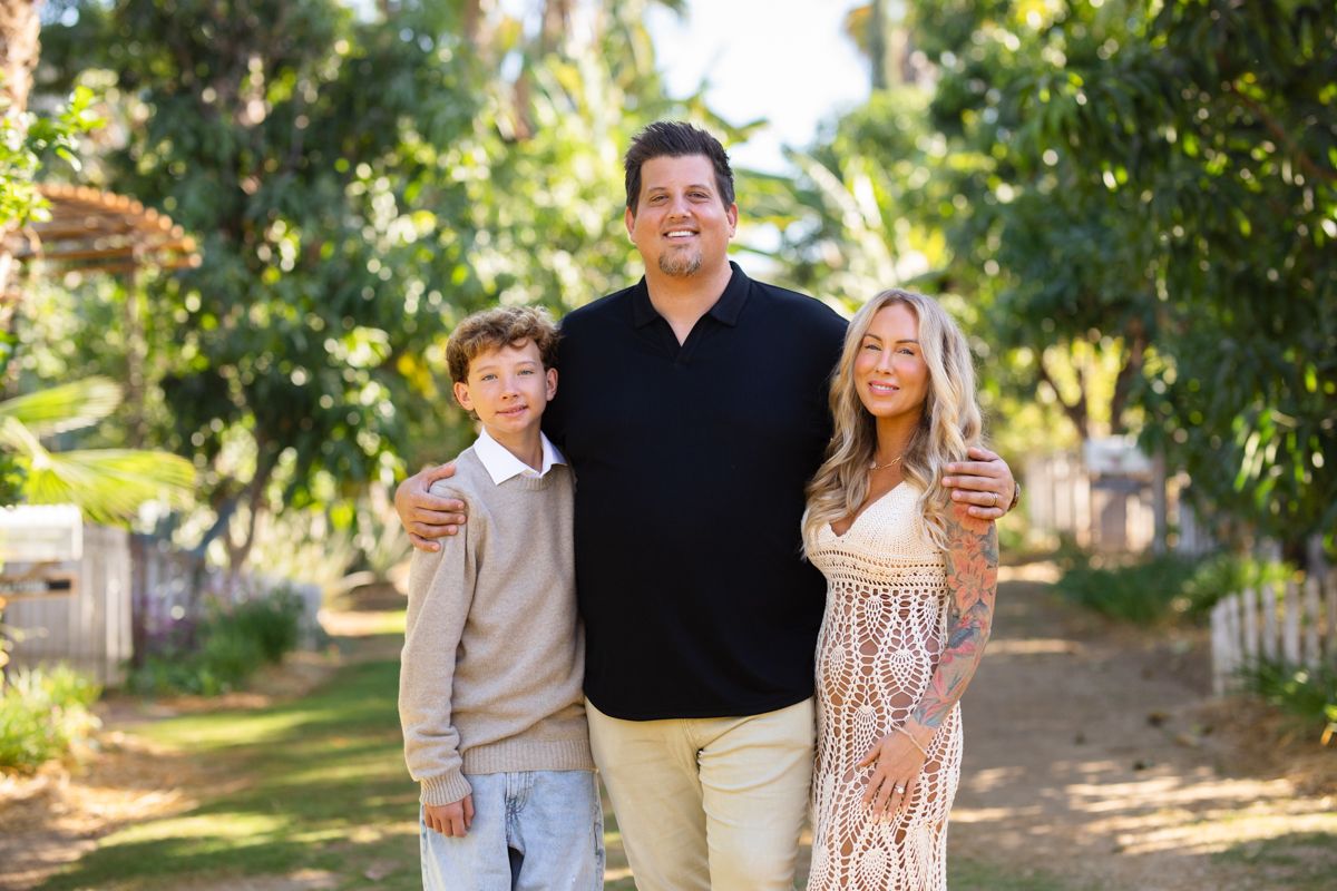 Family of three smiling on a sunny tree-lined path, father with arms around son and daughter.