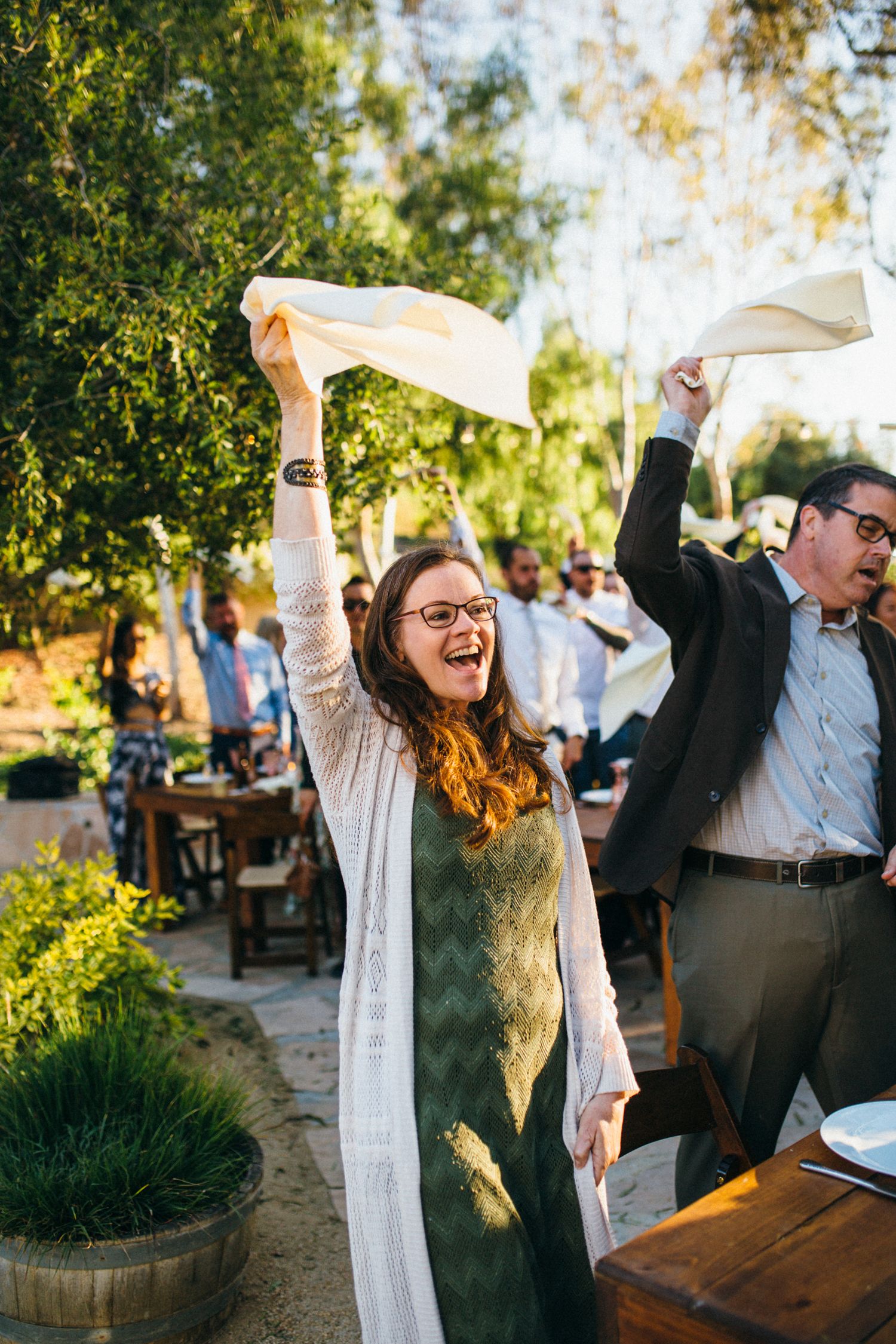 Joyful woman in green dress waves napkin at outdoor wedding reception celebration.