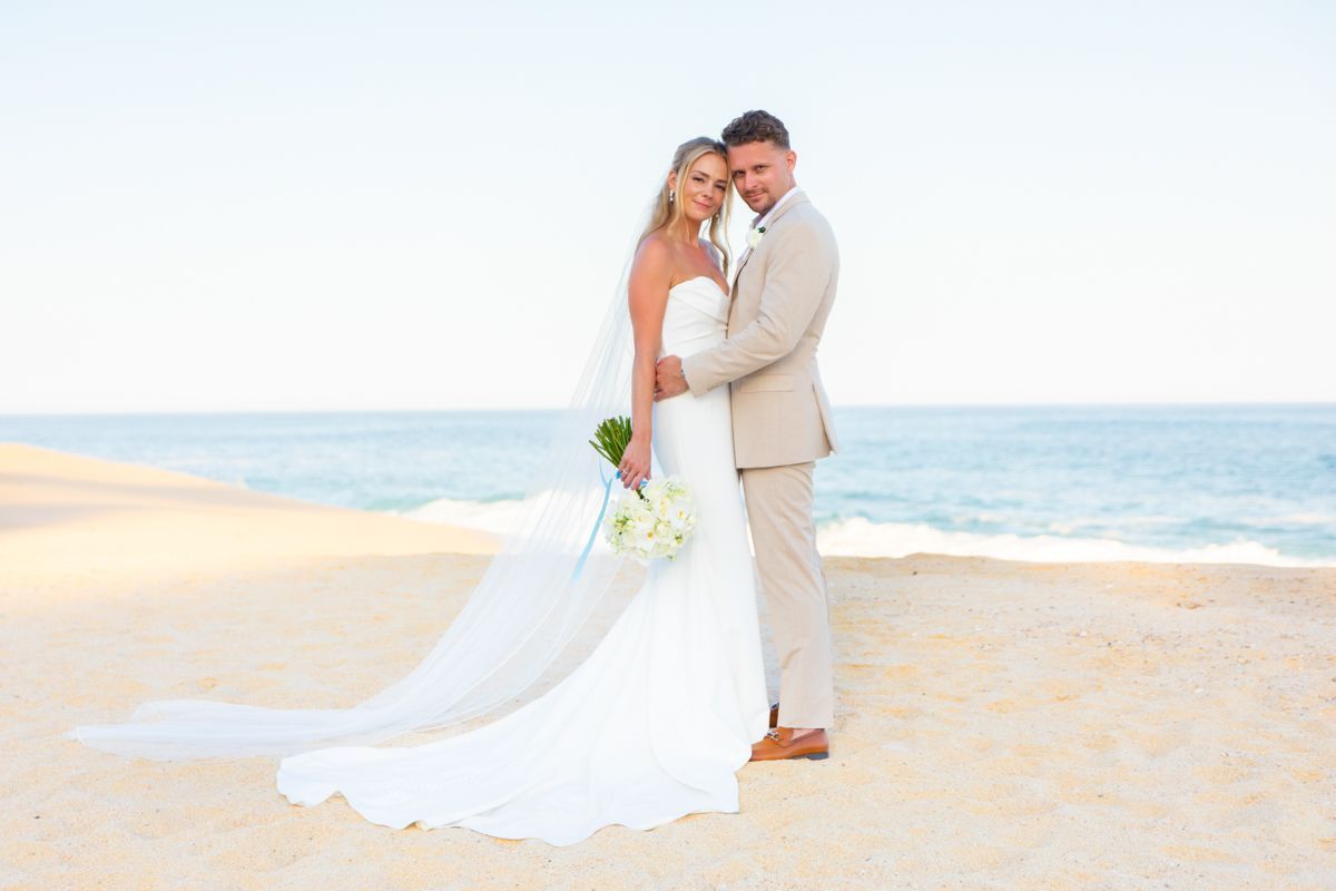 Bride and groom embrace on sandy beach with ocean backdrop in elegant wedding attire.