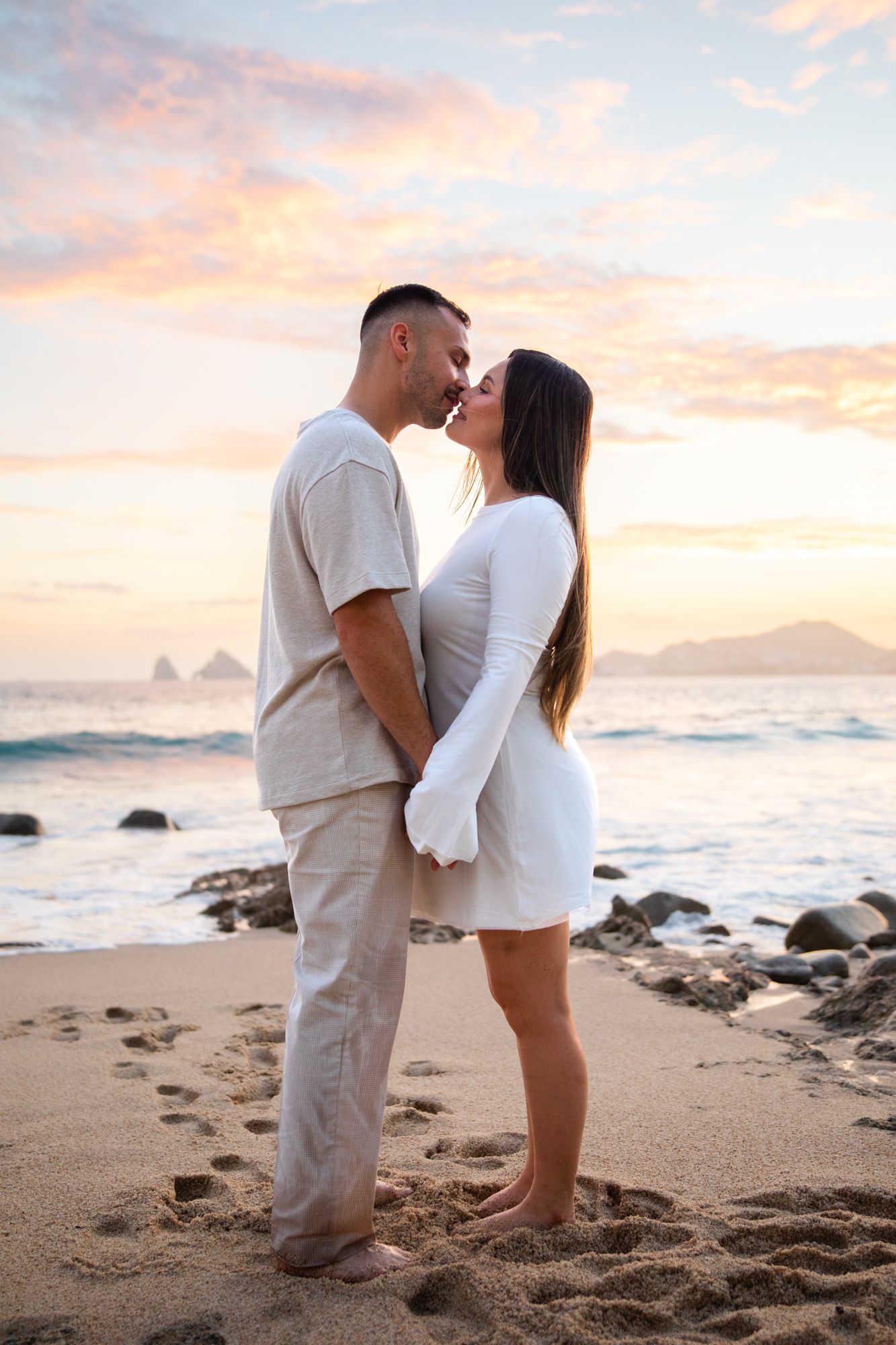 Couple sharing a romantic moment on a sandy beach at sunset, with rocky shores and colorful sky in the background.